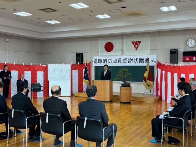 日本の国旗や消防団の旗、紅白幕が飾られた会場で、スーツを着た市長が演台から大勢の人に向かってスピーチをしている様子の写真