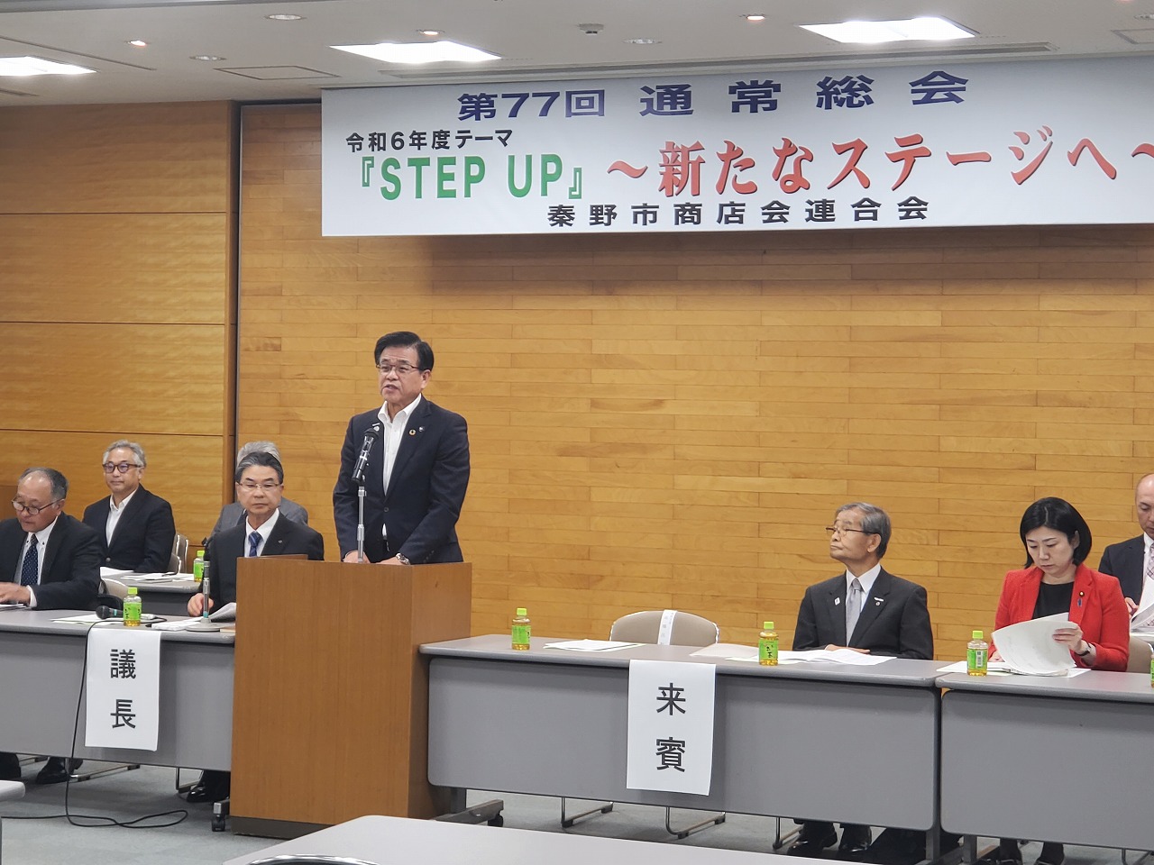 「STEP UP 〜新たなステージへ〜」と書かれた横断幕の下、スーツを着た市長が演台からスピーチをしており、複数の関係者がテーブルに着席している会議の様子の写真