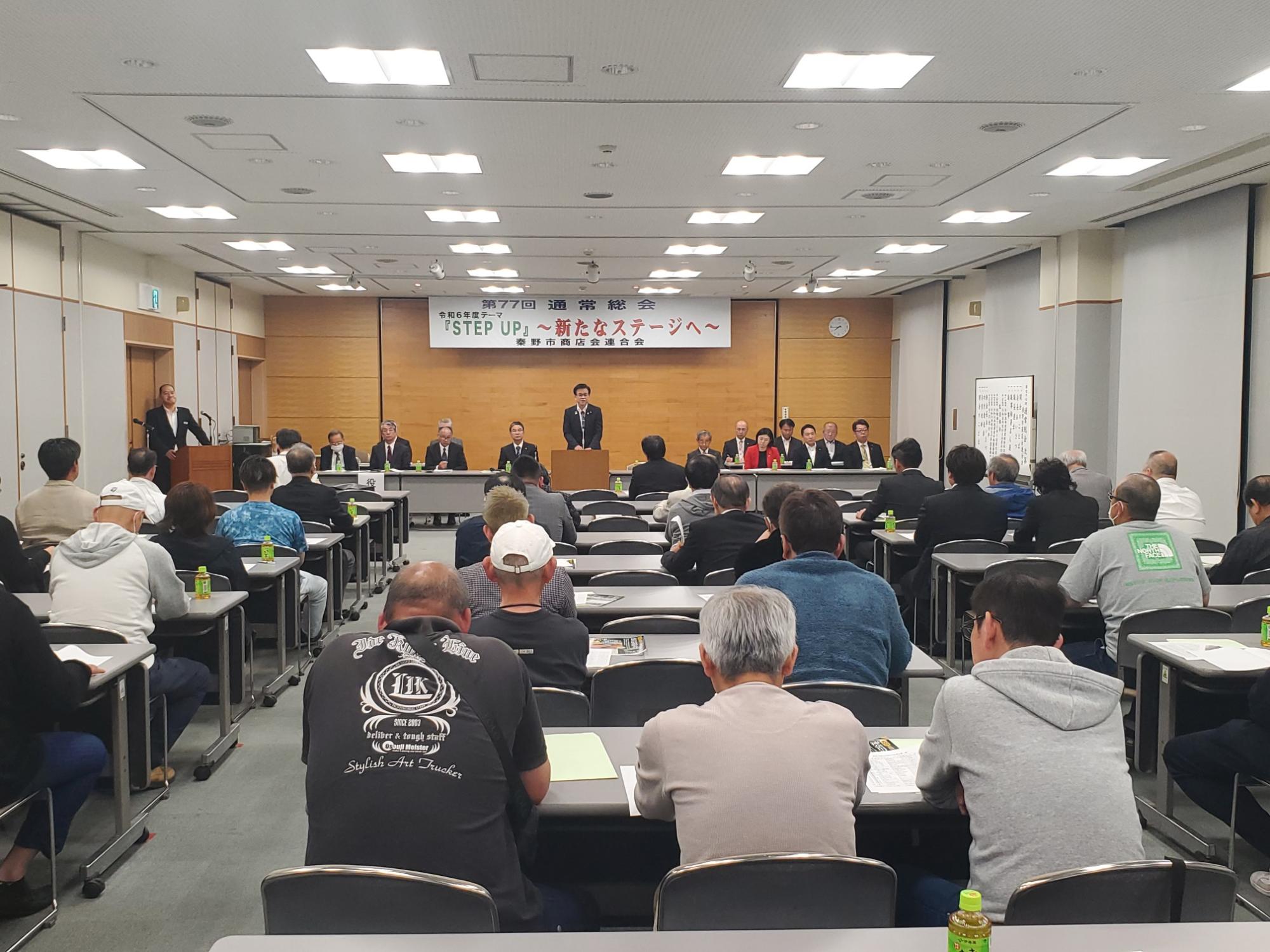 「STEP UP 〜新たなステージへ〜」と書かれた横断幕が掲げられた会場で、スーツ姿の市長が演台から大勢の聴衆や壇上の人々にスピーチをしている様子の写真