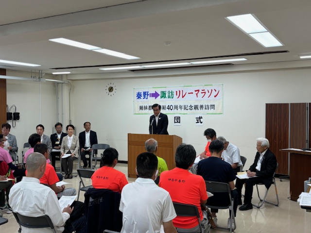 「秦野&rarr;諏訪 リレーマラソン 姉妹都市締結40周年記念親善訪問」の横断幕がある屋内で、スーツ姿の市長が演台からマイクを使ってスピーチをしており、複数の人が椅子に座って話を聞いている様子の写真