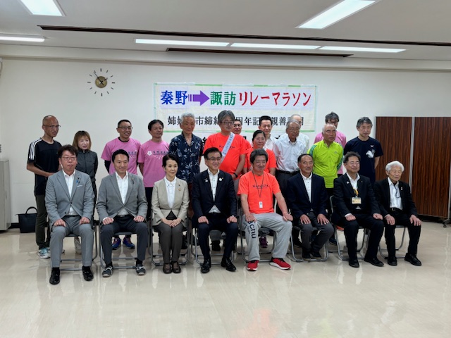 「秦野&rarr;諏訪 リレーマラソン」の横断幕の前で、スーツ姿やマラソンウェアを着た男女のグループが集合写真を撮っている様子の写真