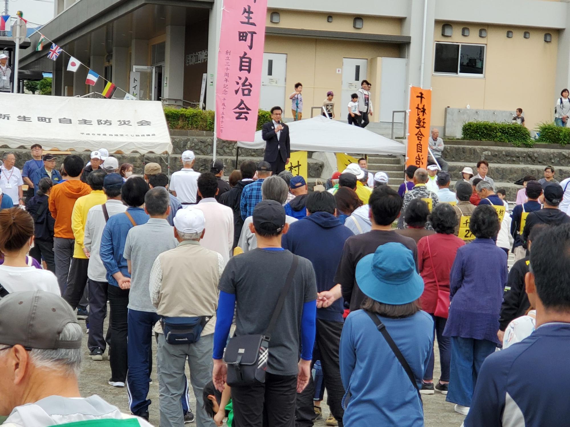 屋外の広場で、スーツ姿の市長がマイクで大勢の聴衆に向かってスピーチをしている様子の写真