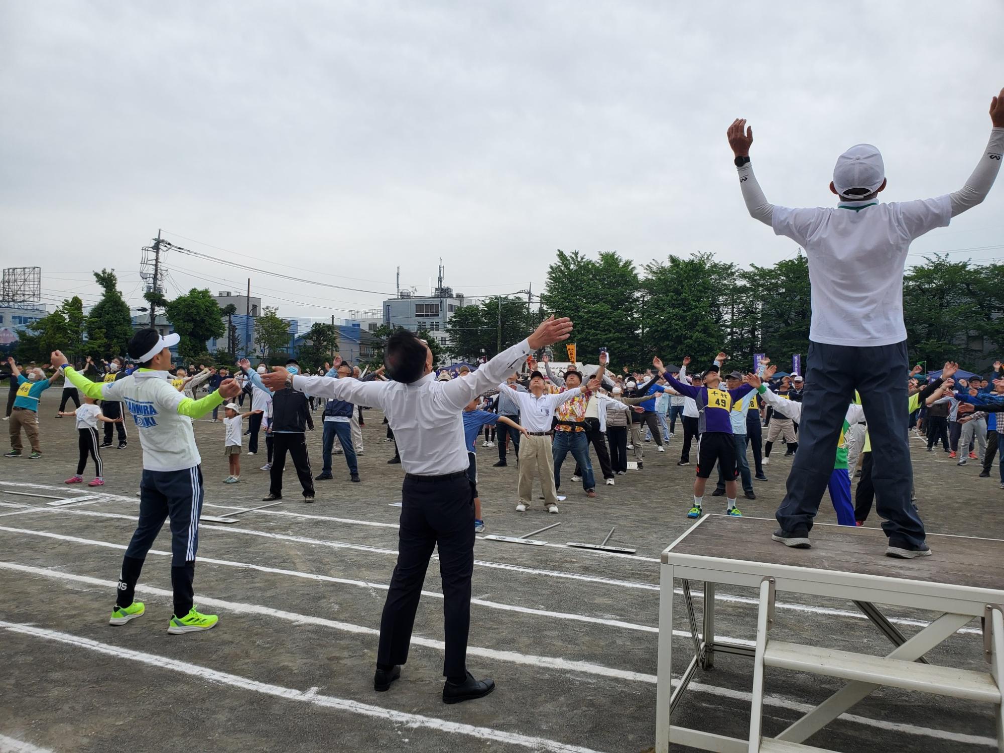 青空の下、大勢の人が運動場で両手を大きく広げた体操をしている様子の写真