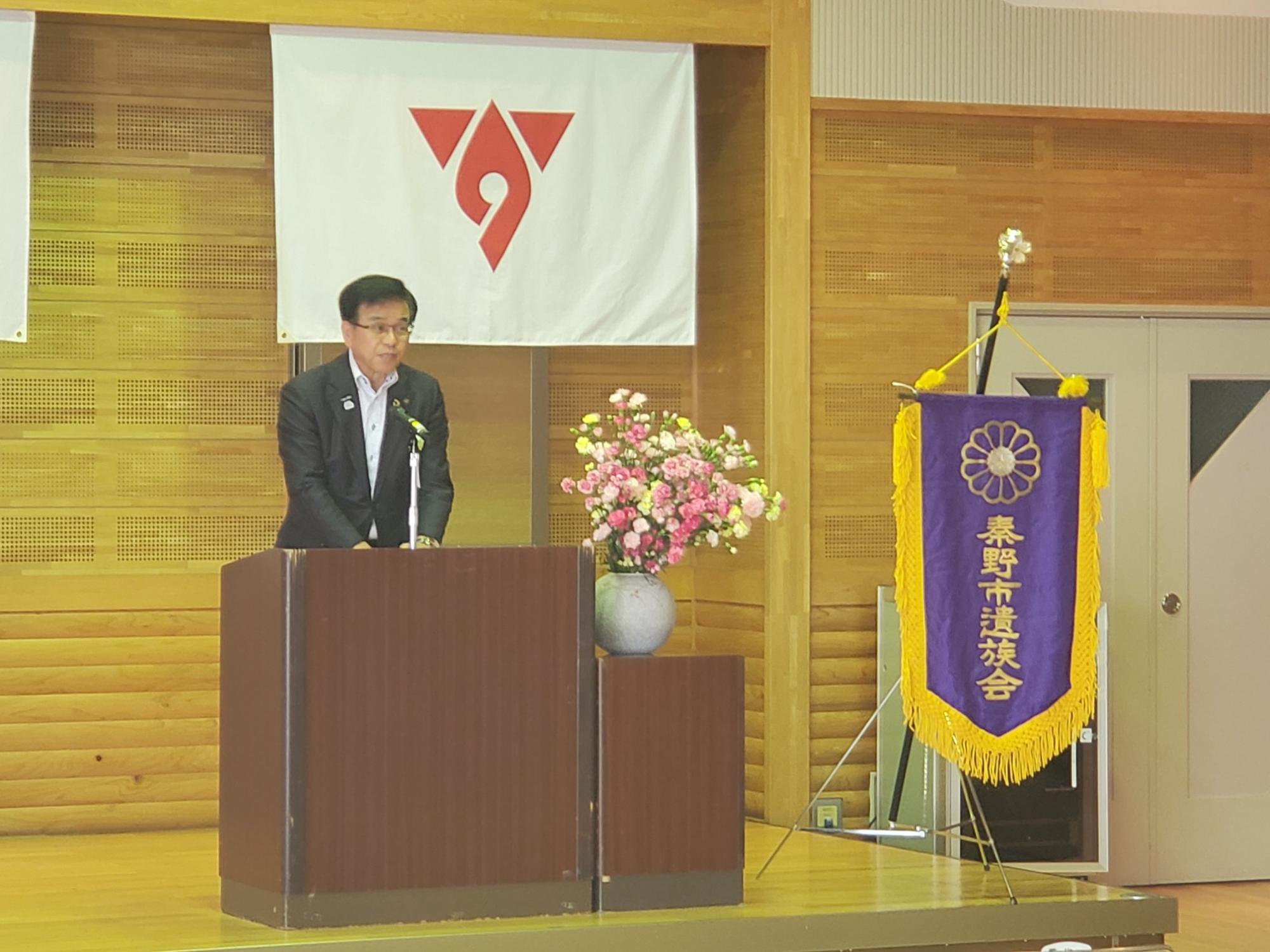 秦野市遺族会と書かれた紫の旗が掲げられた壇上で、スーツ姿の市長がマイクのある演台に立ってスピーチをしている様子の写真