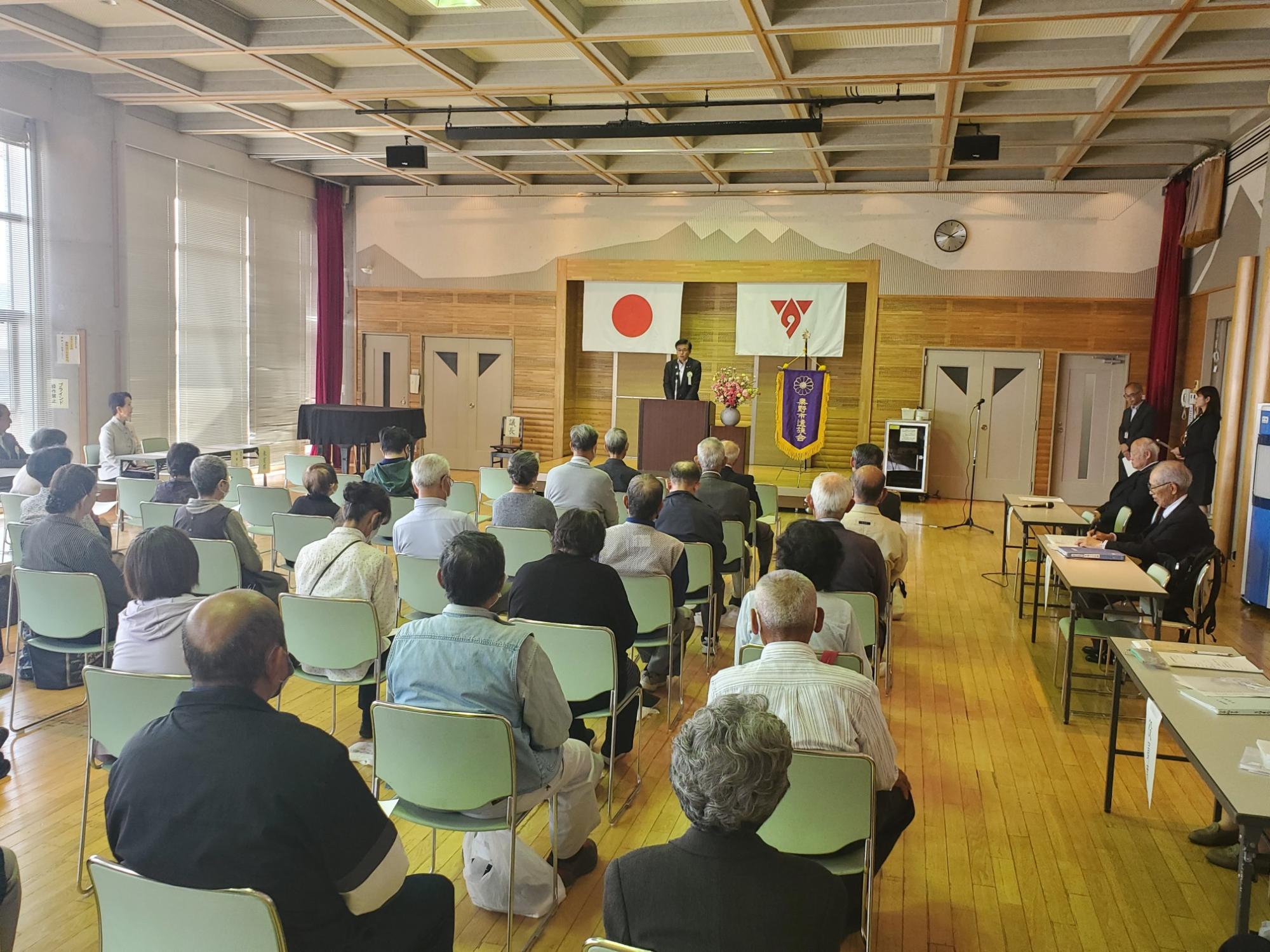 秦野市遺族会と書かれた紫色の旗と日の丸が掲げられた講堂で、スーツ姿の男性が演台に立って大勢の聴衆に向かってスピーチをしている様子の写真