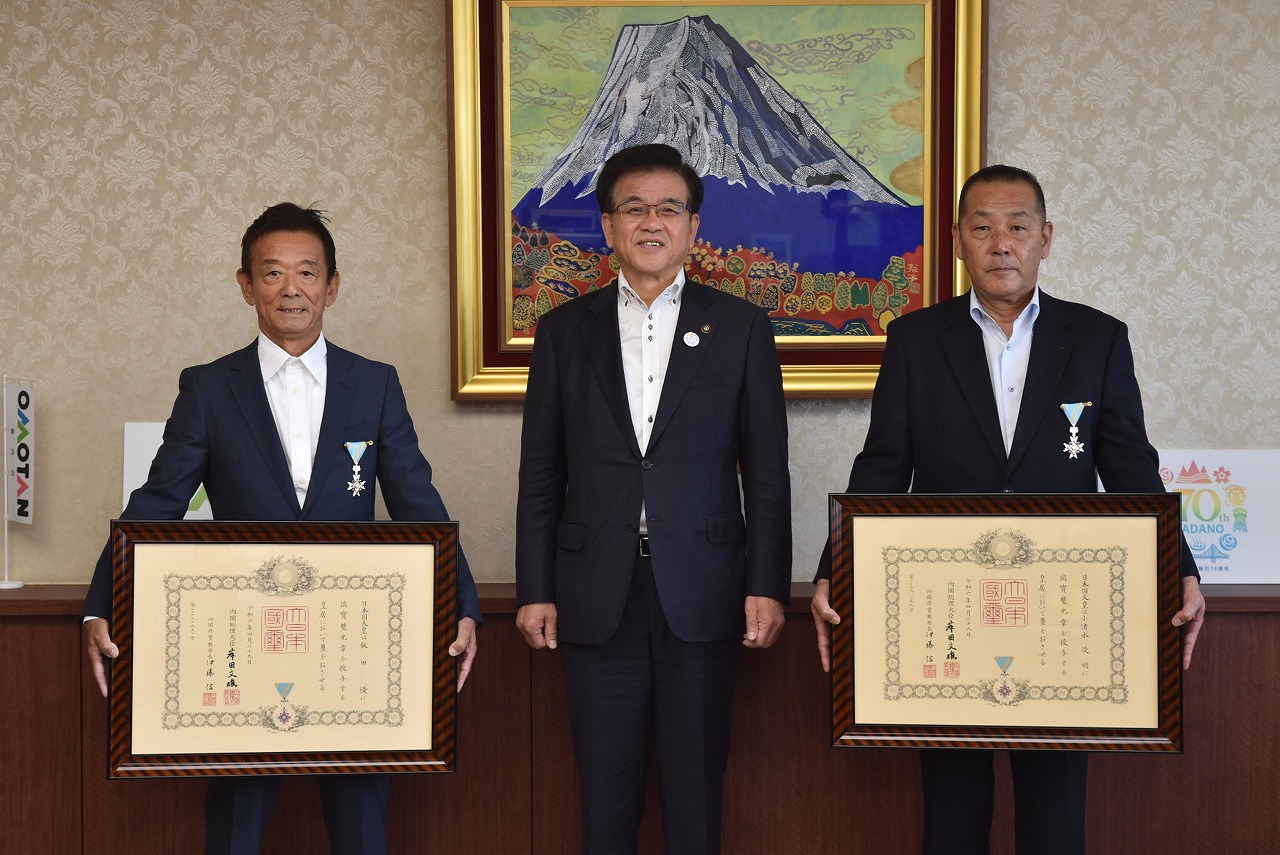 壁に飾られた富士山の絵を背景に、スーツ姿の市長と左右の男性がそれぞれ額縁に入った賞状を両手で持っている様子の写真