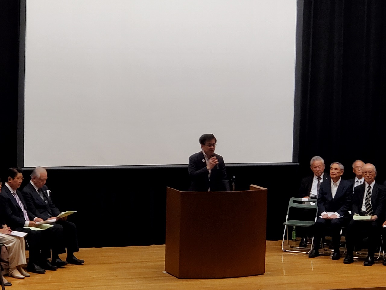 大スクリーンが設置されたステージの演台に立つスーツ姿の市長が、マイクを持ってスピーチしており、その左右にはスーツ姿の関係者が座っている様子の写真