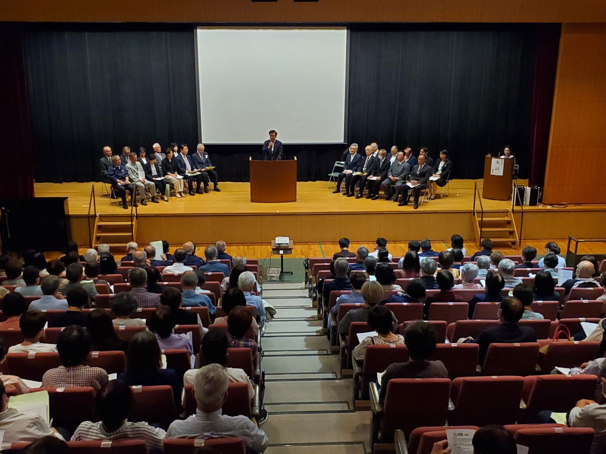 スクリーンと黒いカーテンが背景にあるステージで、スーツ姿の市長が演台から大勢の聴衆に向かってスピーチをしており、壇上には複数の関係者が並んで座っている様子の写真