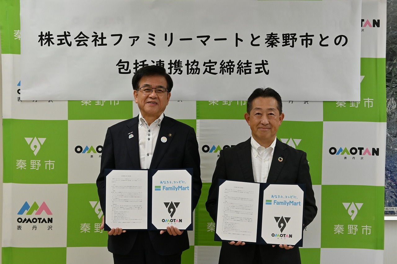 「株式会社ファミリーマートと秦野市との包括連携協定締結式」と書かれた看板の前で、スーツを着た市長と男性がそれぞれ締結書を持ってカメラに向かって立っている様子の写真