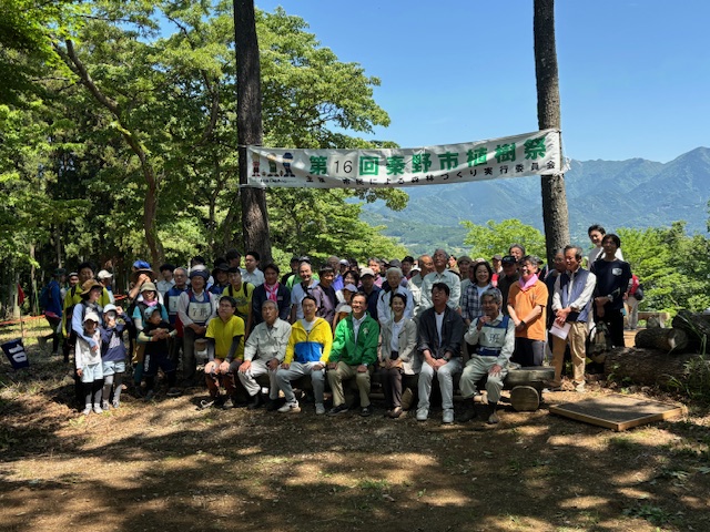 青空の下、山の中で「第16回秦野市植樹祭」と書かれた横断幕を背景に、参加者たちが木製のベンチに座ったり立ったりして集合写真を撮っている様子の写真