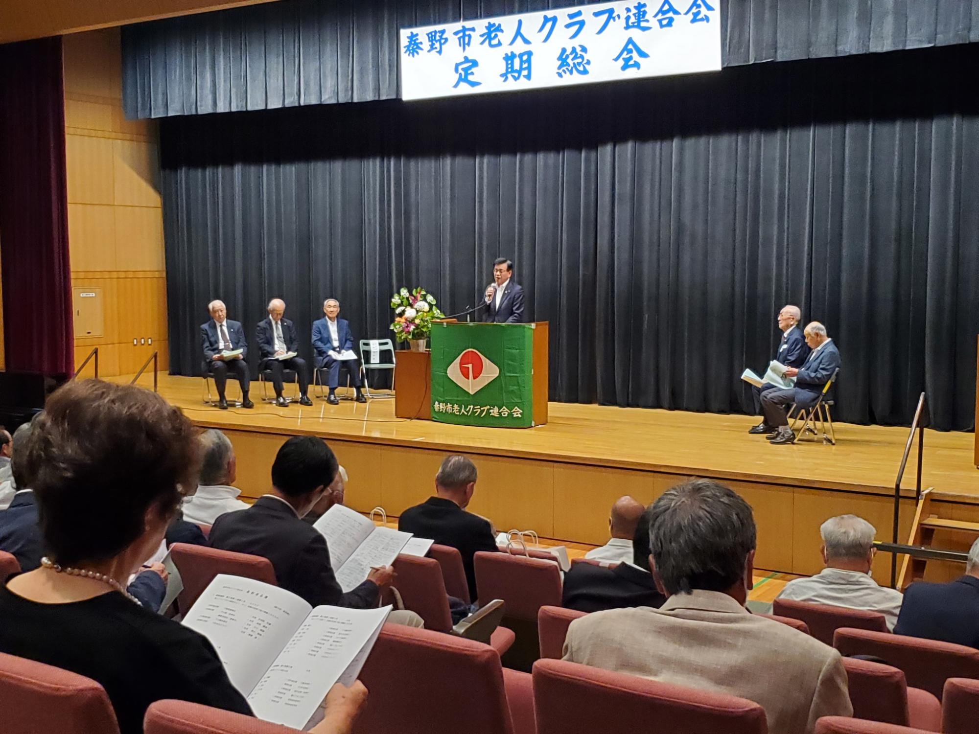 「秦野市老人クラブ連合会 定期総会」と書かれた横断幕の下、スーツ姿の市長が演台からマイクでスピーチをしており、複数の役員が壇上に座り、大勢の参加者が熱心に話を聞いている様子の写真
