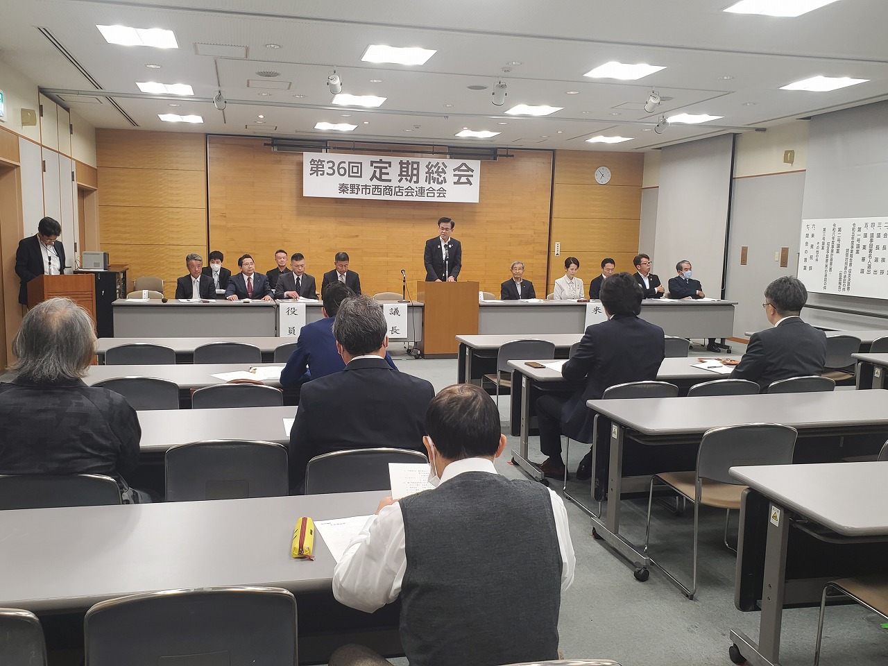 「第36回定期総会 秦野市西商店会連合会」の横断幕が掲げられた会場で、スーツを着た市長が演台からスピーチをしており、複数の参加者が椅子に座って聞いている様子の写真