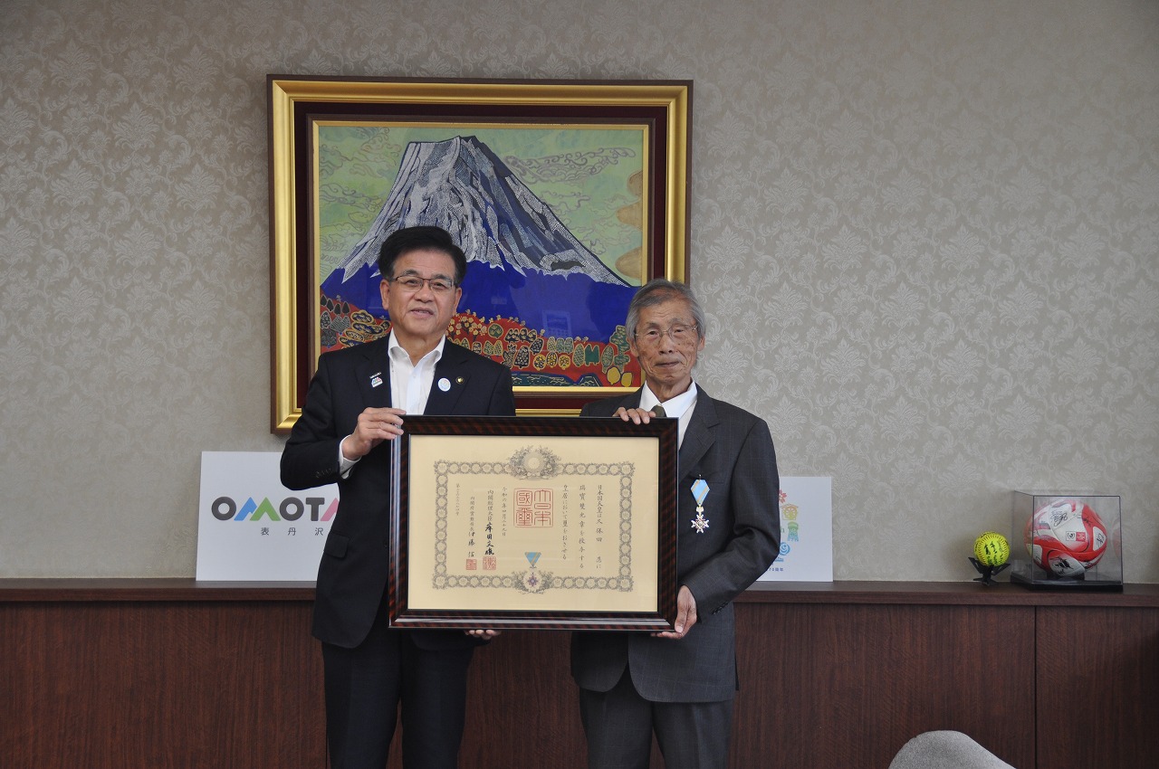 壁に飾られた富士山の絵を背景に、スーツを着た市長と久保田忍さんが額縁に入った賞状を並んで持っている様子の写真