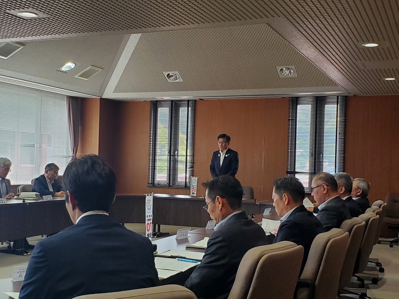 会議室で市長が立って発言し、周囲の委員や職員が資料を見ながら着席して聞いている様子の写真