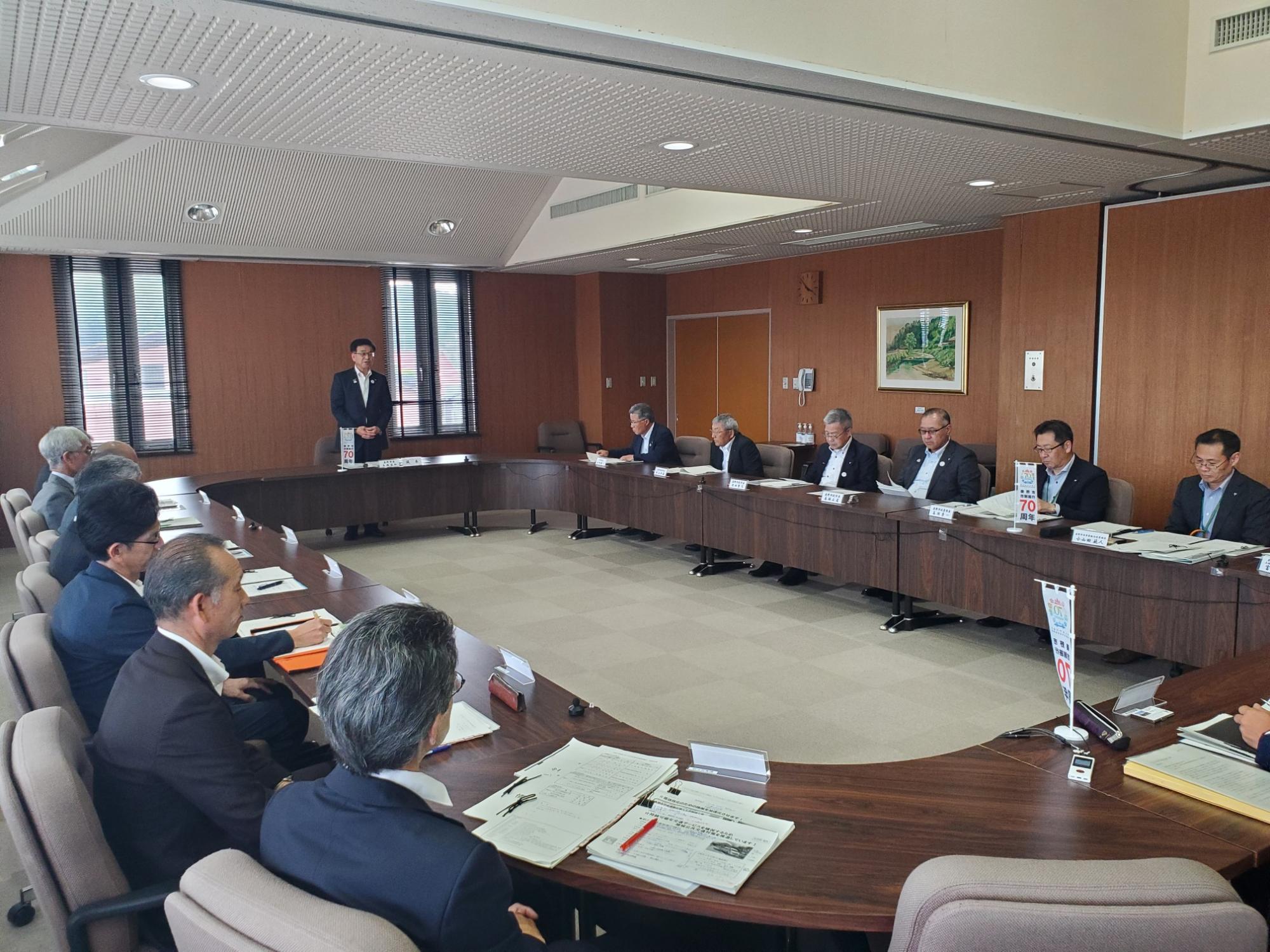会議室で市長がU字型の長机の向こう側に立って挨拶し、名札と資料を手元に置いた十数名の委員や幹部が着席している様子の写真