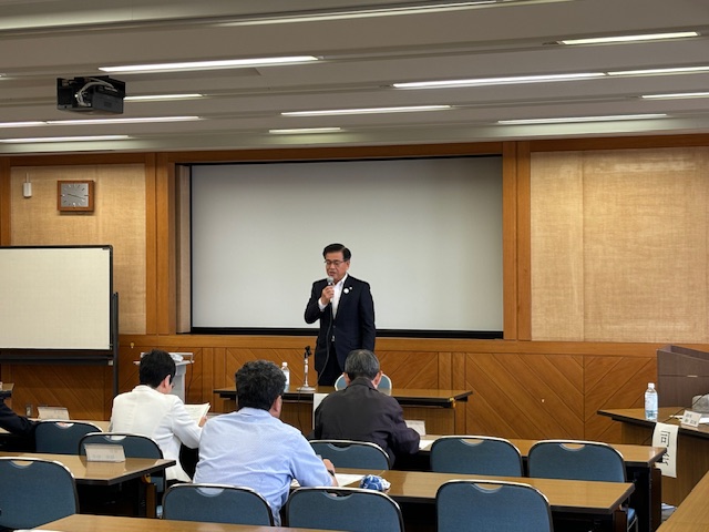 壇上で、スーツ姿の市長がマイクを持ってスピーチしており、その下の机には数名の参加者が座って話を聞いている様子の写真
