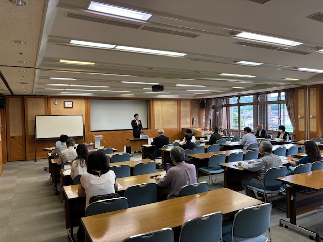 スーツを着た市長が壇上でマイクを持って話し、複数の聴衆が話を聞いている様子の写真