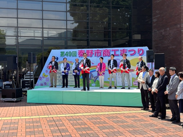 「第49回秦野市商工まつり」の看板がある屋外ステージ上で、法被やスーツを着た人々が横一列に並び、市長が挨拶をしている様子の写真