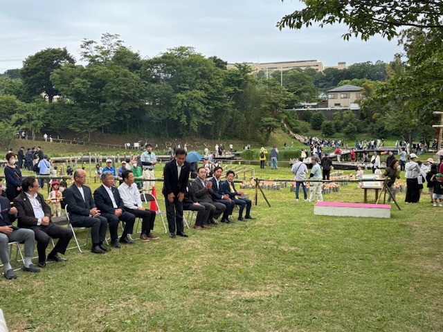 芝生の上で椅子に座る関係者達の列の中央で市長が立って頭を下げており、奥の池の周りで多くの人々が活動している様子の写真