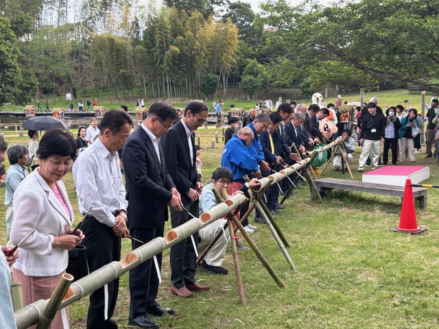 緑豊かな広場で、多くの人々が竹の筒に点灯の準備をする様子の写真