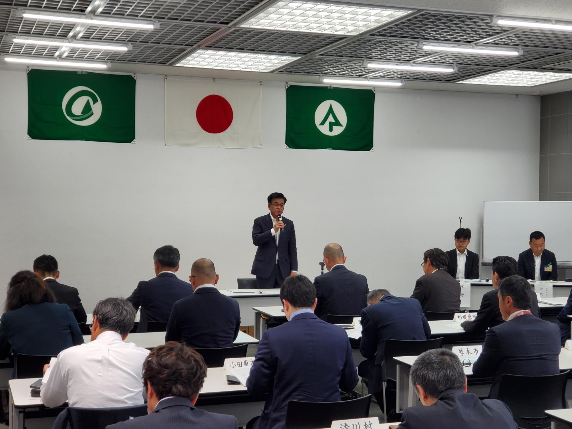 2つの緑の旗と日の丸が掲げられた部屋の前に、スーツを着た市長が演台に立ち、大勢の参加者に向かってマイクでスピーチをしている様子の写真