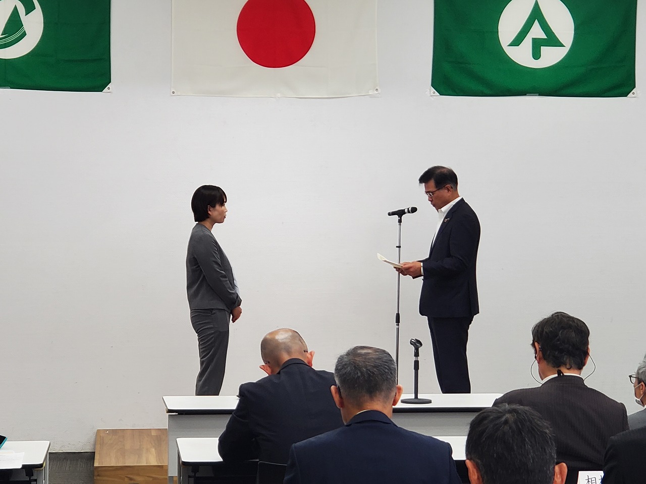 白い壁を背景に日の丸と緑の旗が掲げられた部屋で、スーツ姿の男性がマイクスタンドの前で女性に賞状を渡している様子の写真