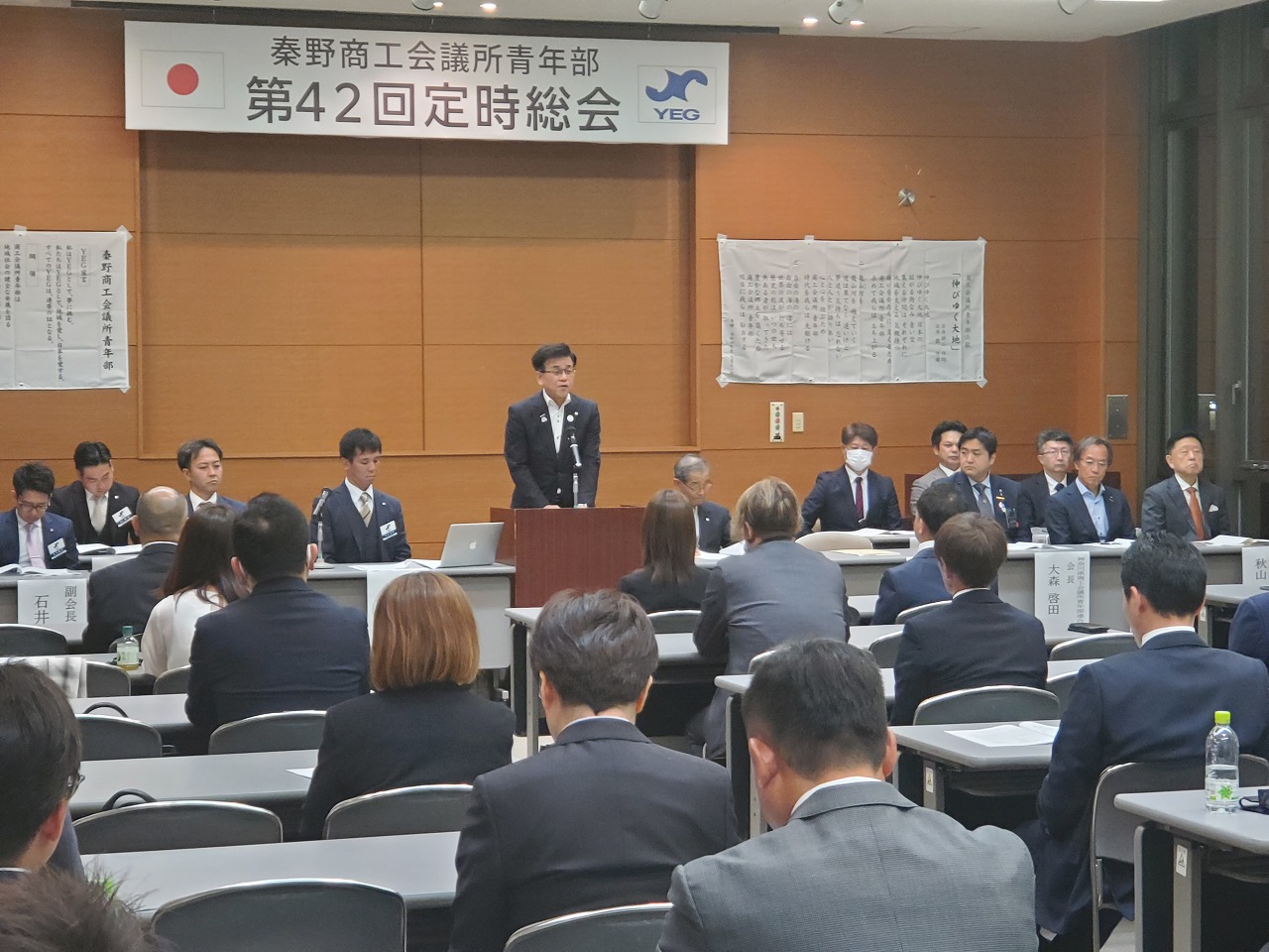「秦野商工会議所青年部 第42回定時総会」と書かれた横断幕の下で、スーツを着た市長が演台に立ち、大勢の聴衆や壇上の人々にスピーチをしている様子の写真