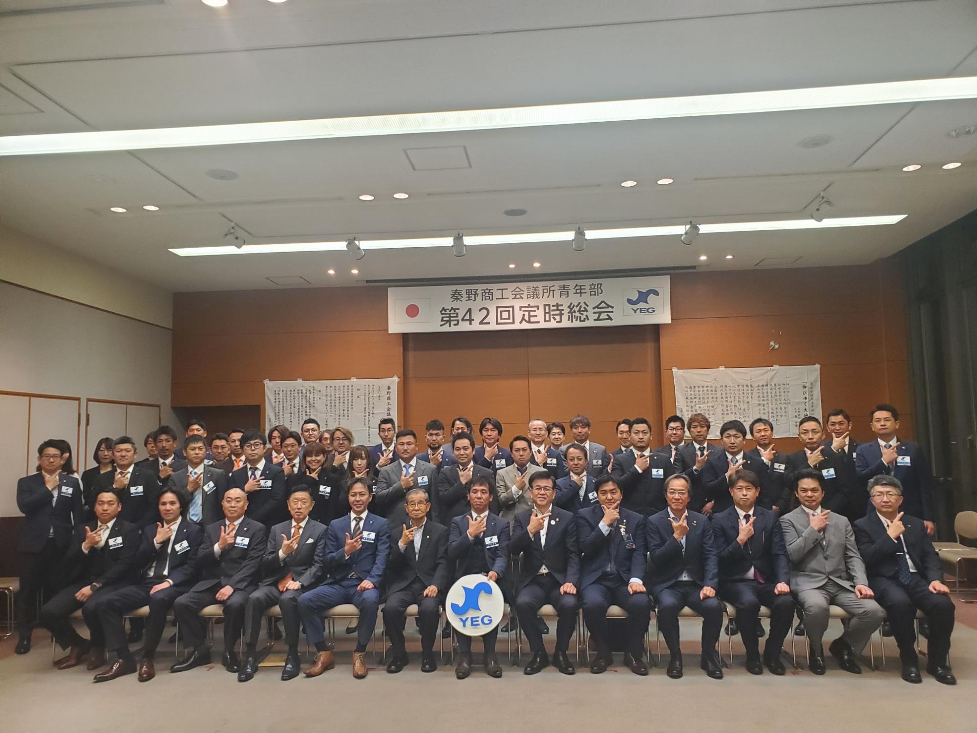 「秦野商工会議所青年部 第42回定時総会」と書かれた横断幕の前で、スーツ姿の複数の男女が一列に座り、その後ろに立った他の人々とともに集合写真を撮っている様子の写真