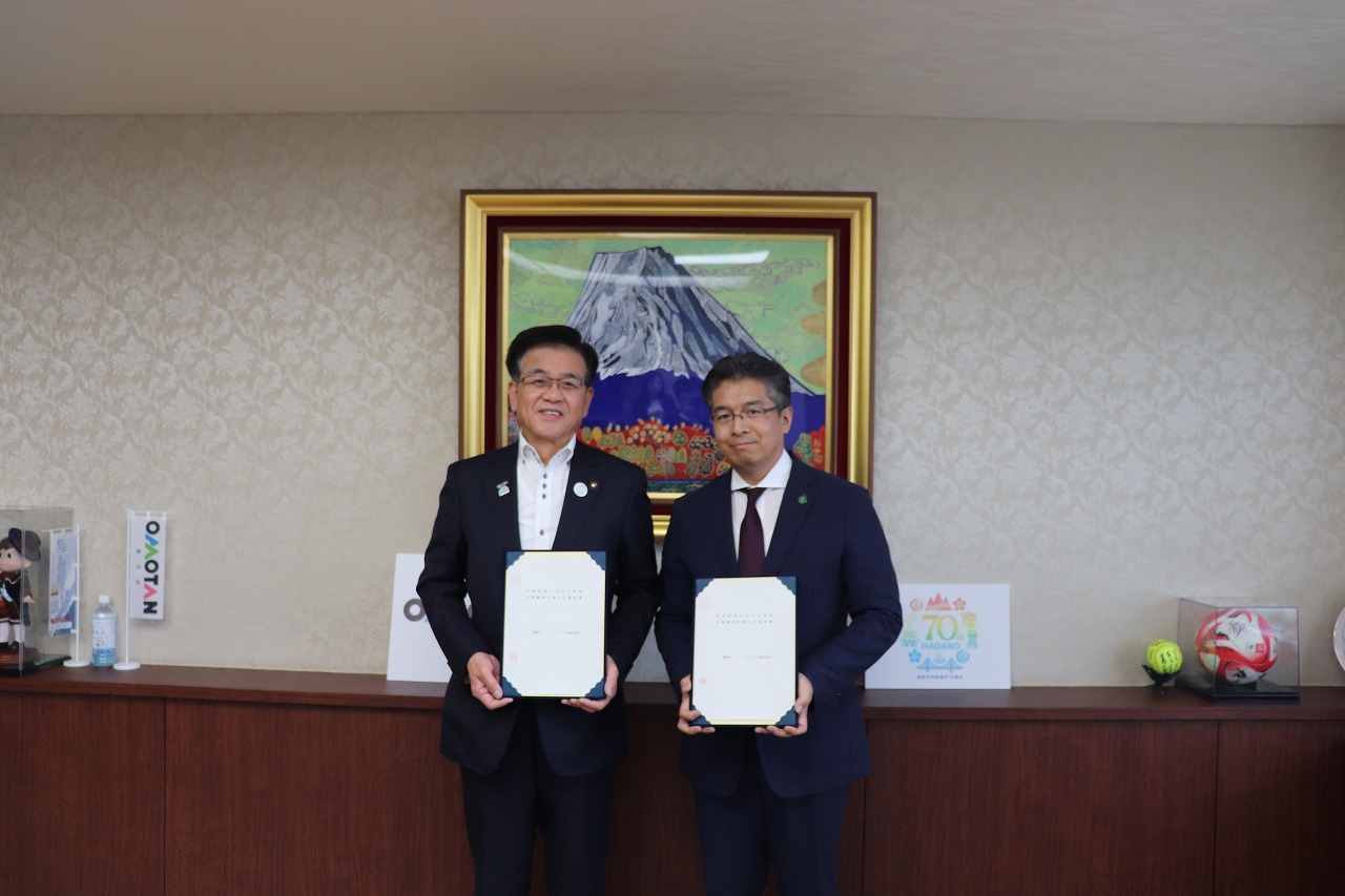 壁に飾られた富士山の絵を背景に、スーツを着た市長と男性が協定書を並んで持っている様子の写真