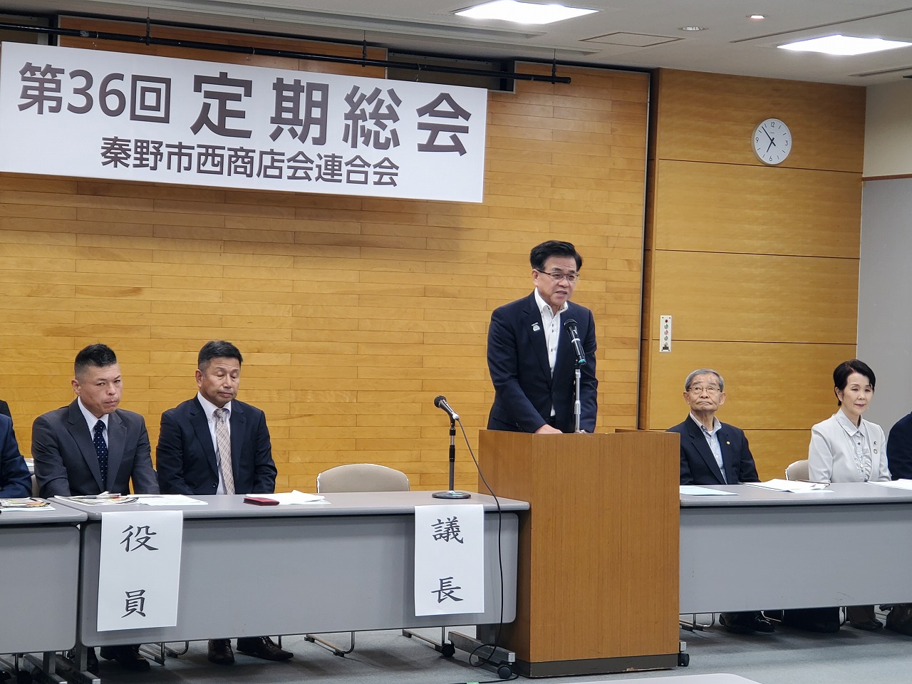 「第36回定期総会 秦野市西商店会連合会」と書かれた横断幕がある会場で、スーツを着た市長が演台に立ってスピーチをしており、複数の役員が座って聞いている様子の写真
