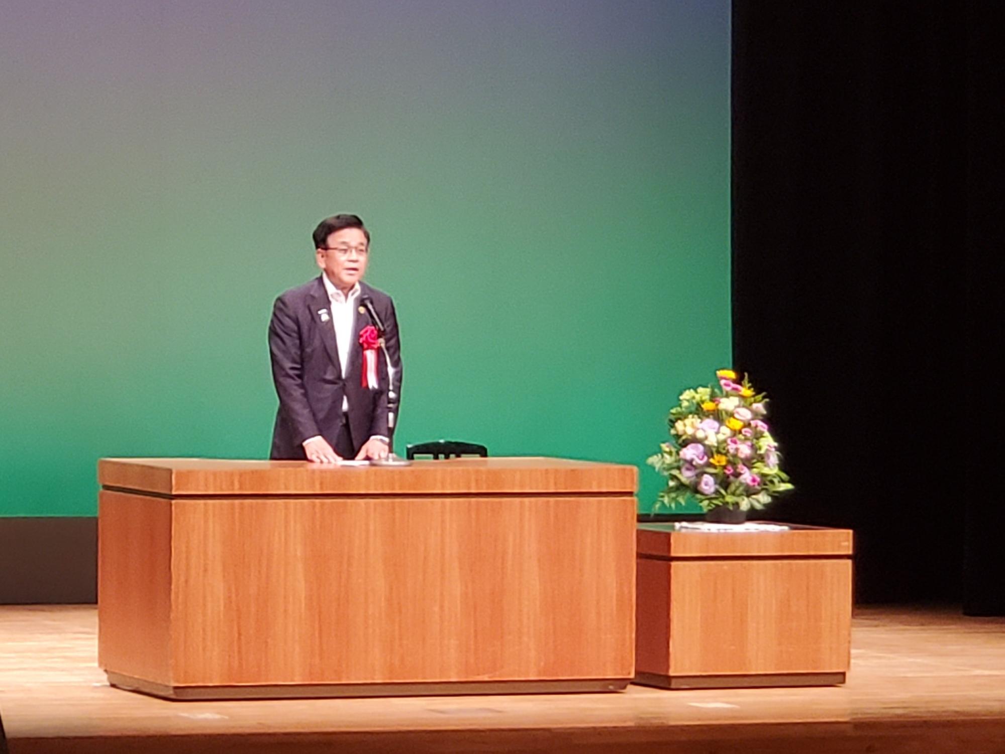 薄い緑の背景のステージで、スーツを着て左胸に赤いリボンのついた市長が、木製の演台から身振り手振りを交えながらスピーチしている様子の写真