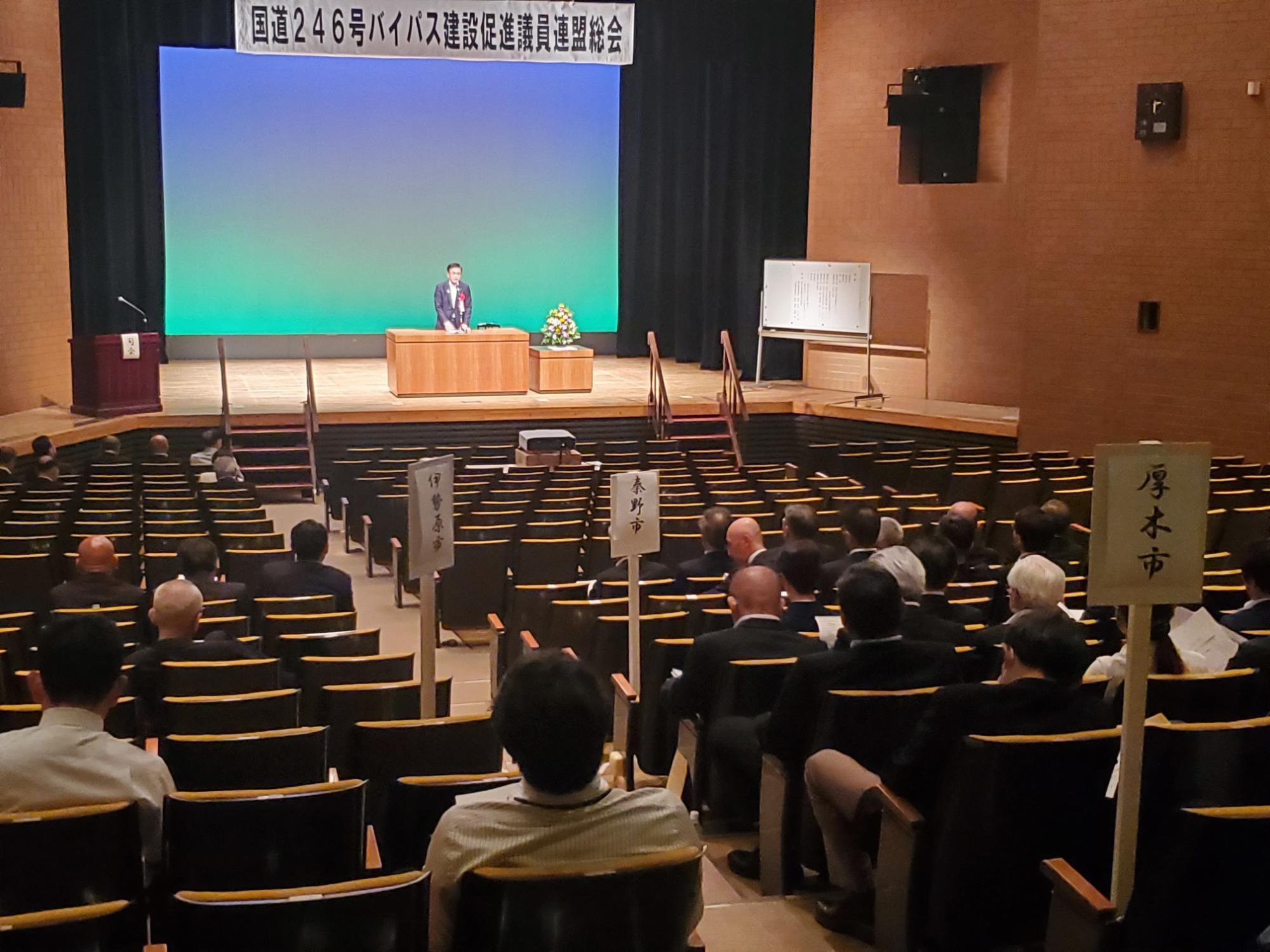 「国道246号バイパス建設促進議員連盟総会」と書かれた看板の下、スーツ姿の市長がマイクのある演台から大勢の聴衆に向かってスピーチしている様子の写真