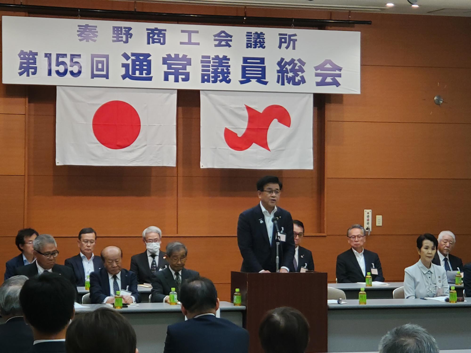 「秦野商工会議所 第155回通常議員総会」と書かれた横断幕と日の丸の下、スーツ姿の市長が演台からスピーチをしており、壇上には複数の人が着席している会議の様子の写真