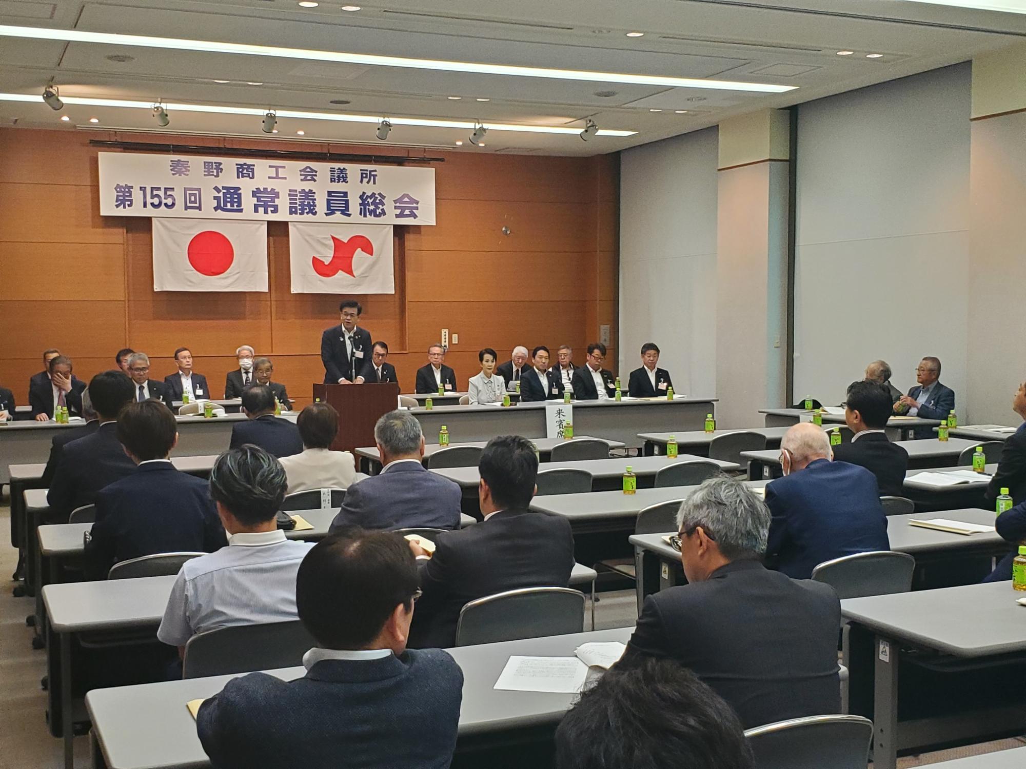 「秦野商工会議所 第155回通常議員総会」と書かれた横断幕と日の丸の下で、スーツ姿の市長が演台から大勢の聴衆に向かってスピーチしている会議の様子の写真