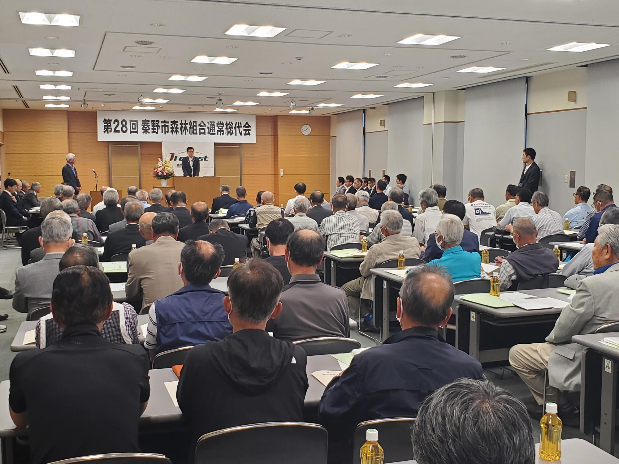 「第28回秦野市森林組合通常総代会」と書かれた横断幕がある会場で、多くの参加者が着席し、スーツを着た市長が演台に立ち、スーツ姿の男性が演台から大勢の聴衆に向かって話している様子の写真