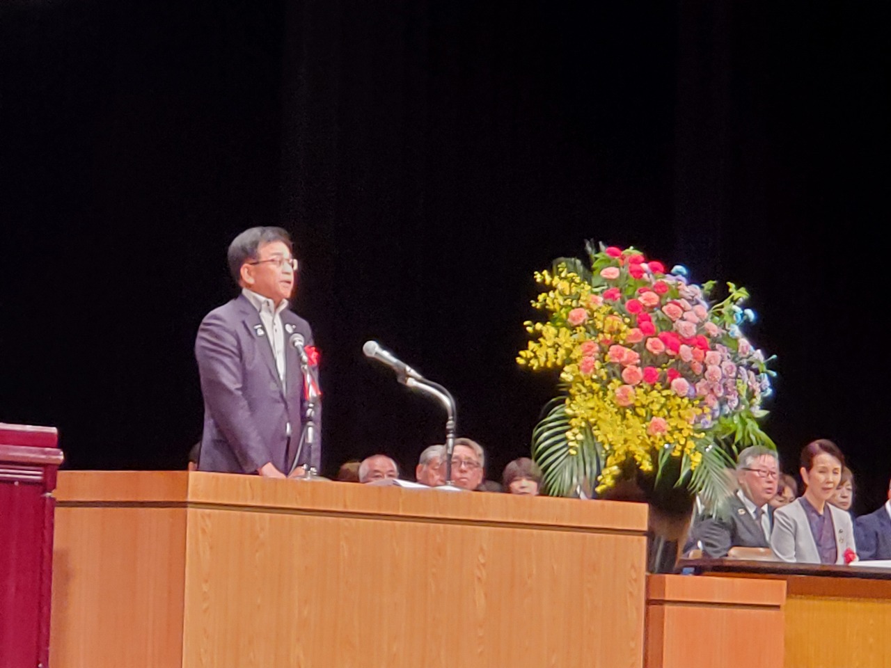 ステージ上の木製演台に立つスーツ姿の市長がマイクに向かってスピーチしており、その横には赤や黄色の花が豪華に飾られた大きな花束が置かれている写真