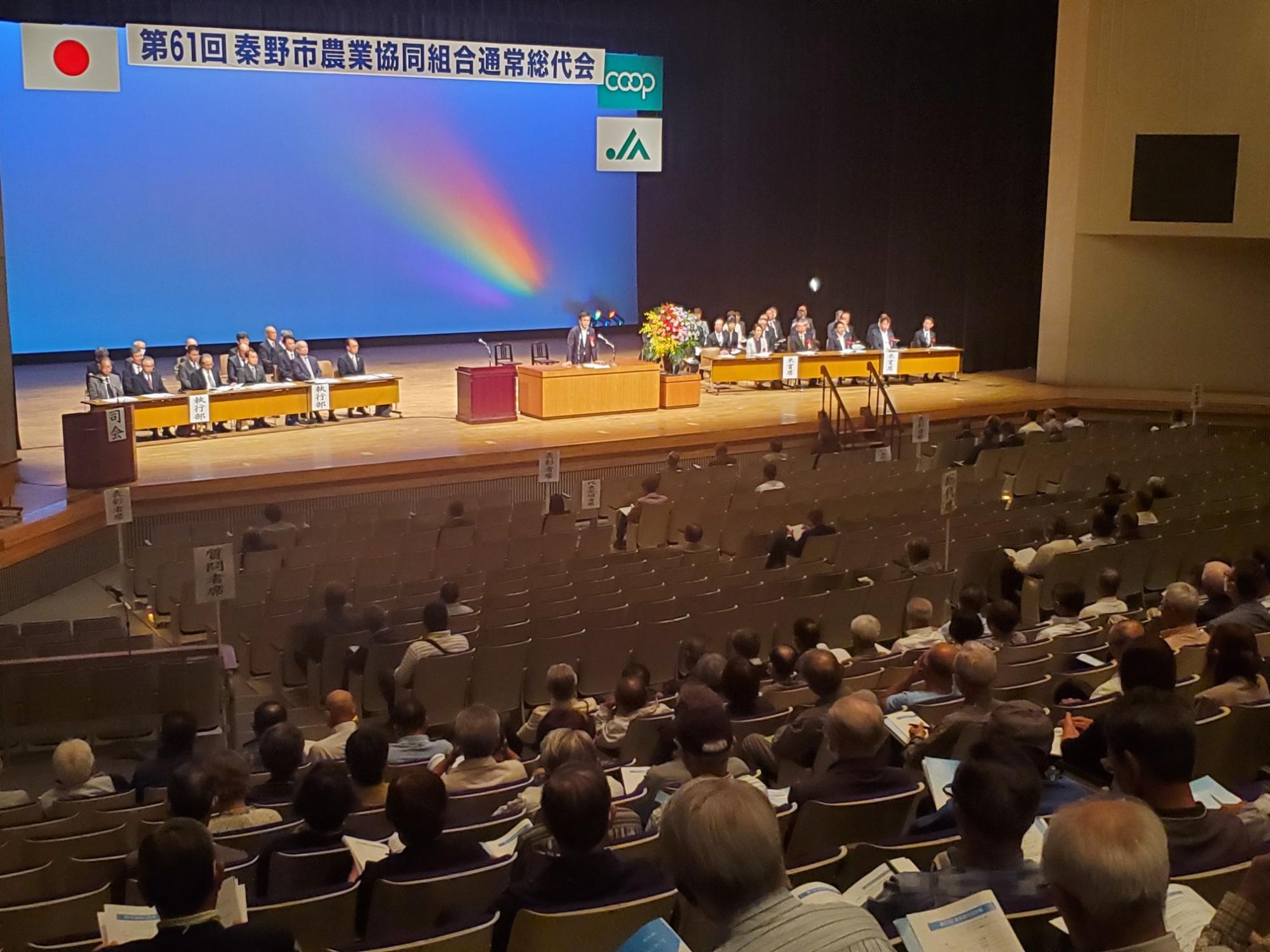「第61回秦野市農業協同組合通常総代会の会場で、ステージ上に関係者が横一列に並んで着席し、演壇に立つ男性が挨拶をしており、客席には多くの総代や関係者が資料を手にして着席している様子の写真
