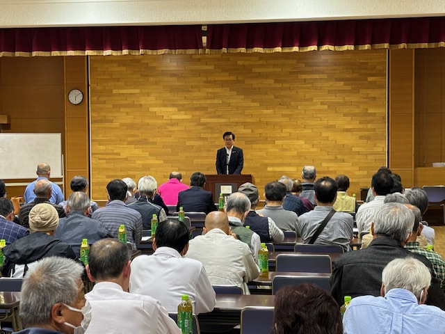 複数のテーブルと椅子が並んだ広間で、スーツ姿の市長が演台から大勢の聴衆に向かってスピーチをしている様子の写真