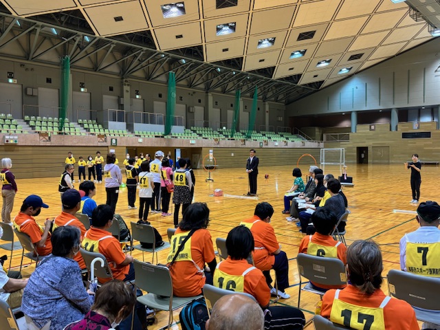 広い屋内体育館で、黄色のビブスを着用した参加者を含む多くの人々が椅子に座ったり立ったりしている中、スーツ姿の市長が中央に立って、スポーツイベントの開会式の挨拶をしている様子の写真