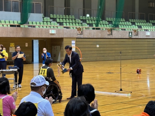 体育館の中で、スーツ姿の市長が車椅子に座った参加者にトロフィーを手渡しており、周囲では複数のスタッフと観客が拍手をして、スポーツイベントの表彰式が行われている様子の写真