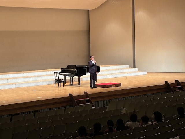 茶色の木製床の舞台に黒いグランドピアノが置かれ、その前に立つスーツ姿の市長が客席に向かってマイクで挨拶をしており、手前の客席には数名の聴衆の頭部が見える、コンサートホールでのイベントの様子の写真