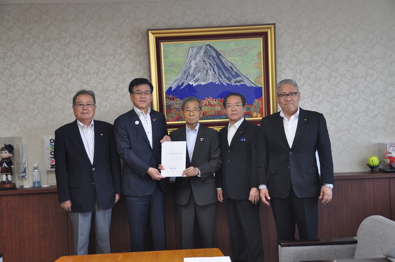 富士山の絵画が壁に飾られた部屋で、スーツ姿の市長と四人の男性が一列に並び、中央の二人が白い書類の束を持って写真に収まっている写真