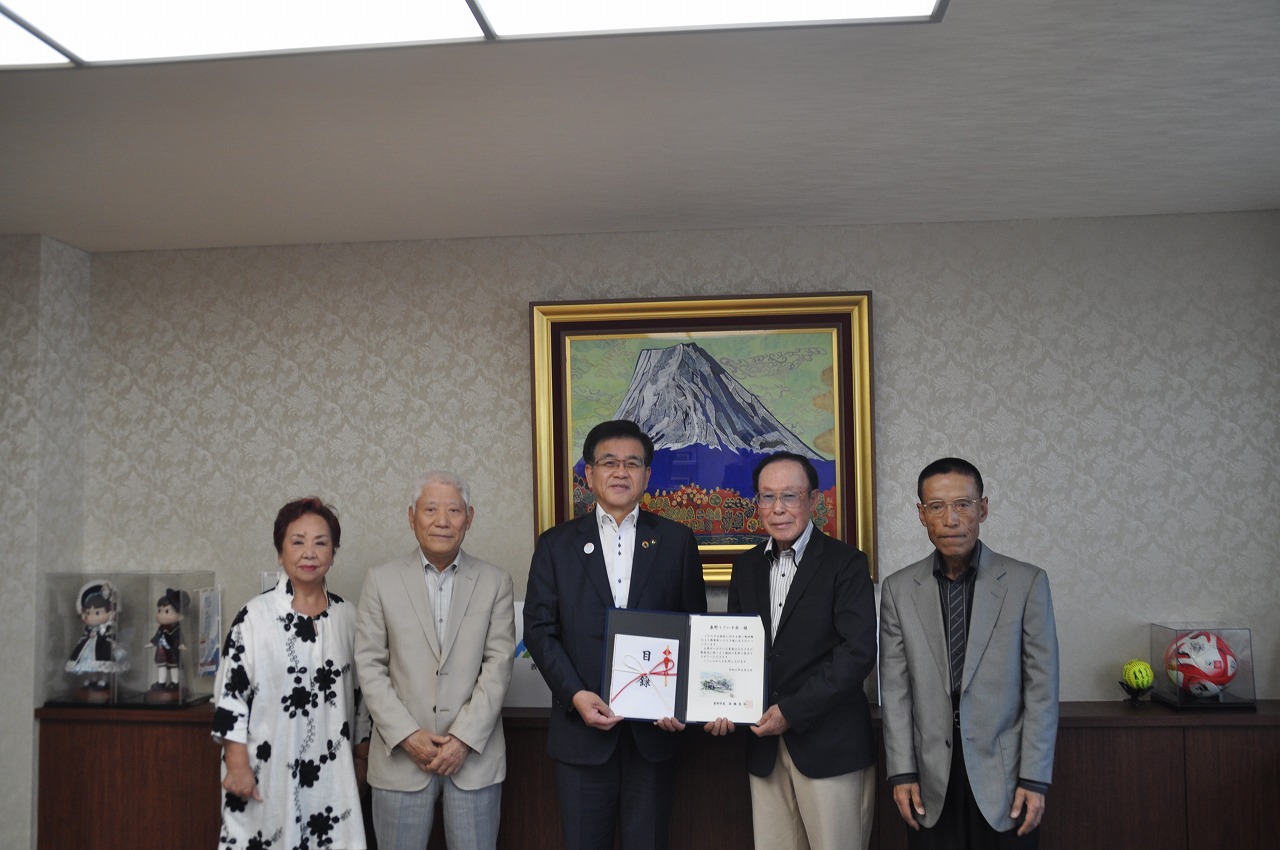 富士山の絵画が飾られた部屋で、スーツ姿の市長を中心に、右手に賞状のようなものを持った男性、左手には明るい色のスーツ姿の男性と白黒模様のワンピースを着た女性、そして右端に濃いスーツを着た男性が一列に並んで立つ集合写真