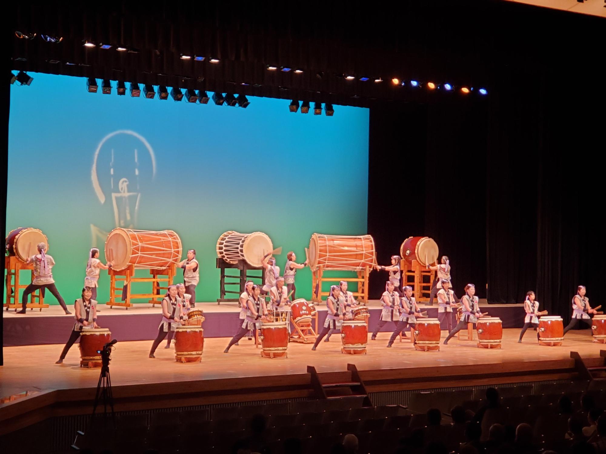 舞台上の複数の奏者が、大きさの異なる多数の和太鼓を前に、青い照明が当たる背景幕の中央に白いロゴが浮かび上がる演出のもと、客席に向かって一列に並んで演奏している迫力ある舞台風景の写真