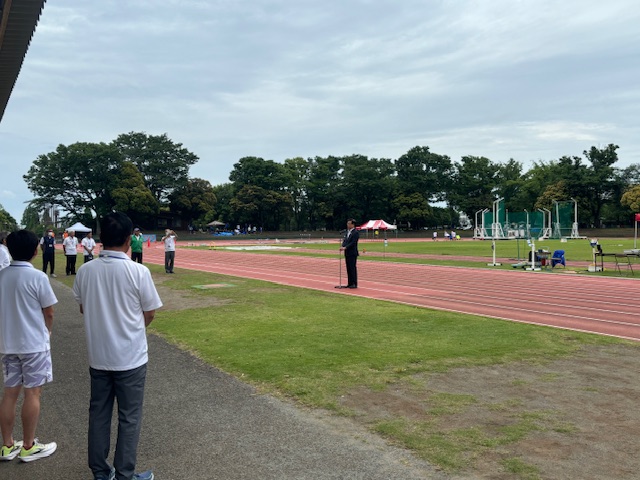 遠景には木々と投てき用の防護ネットが見える運動公園の陸上競技場で、数人の聴衆が距離を空けて立ち並び、トラックに面した芝生の上でスーツ姿の市長がマイクを持って挨拶をしている開会式の写真