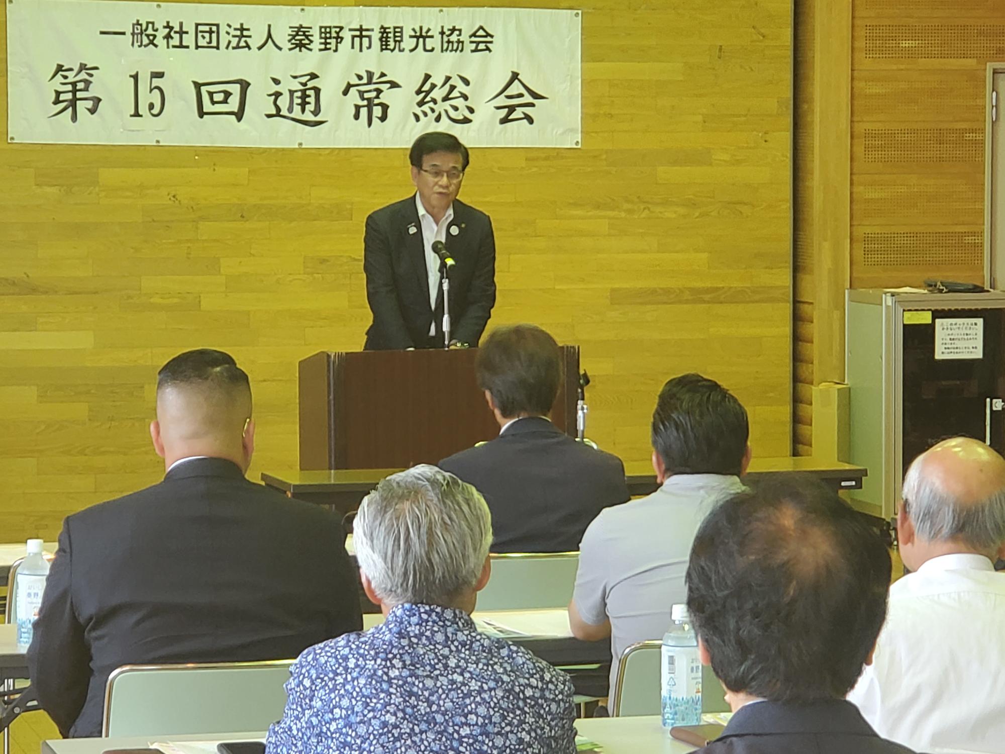 市長が、木目調の壁を背景に「一般社団法人秦野市観光協会 第15回通常総会」と書かれた横断幕の下、聴衆に向かって演台でマイクを使って話している様子を参加者の後方から捉えた写真
