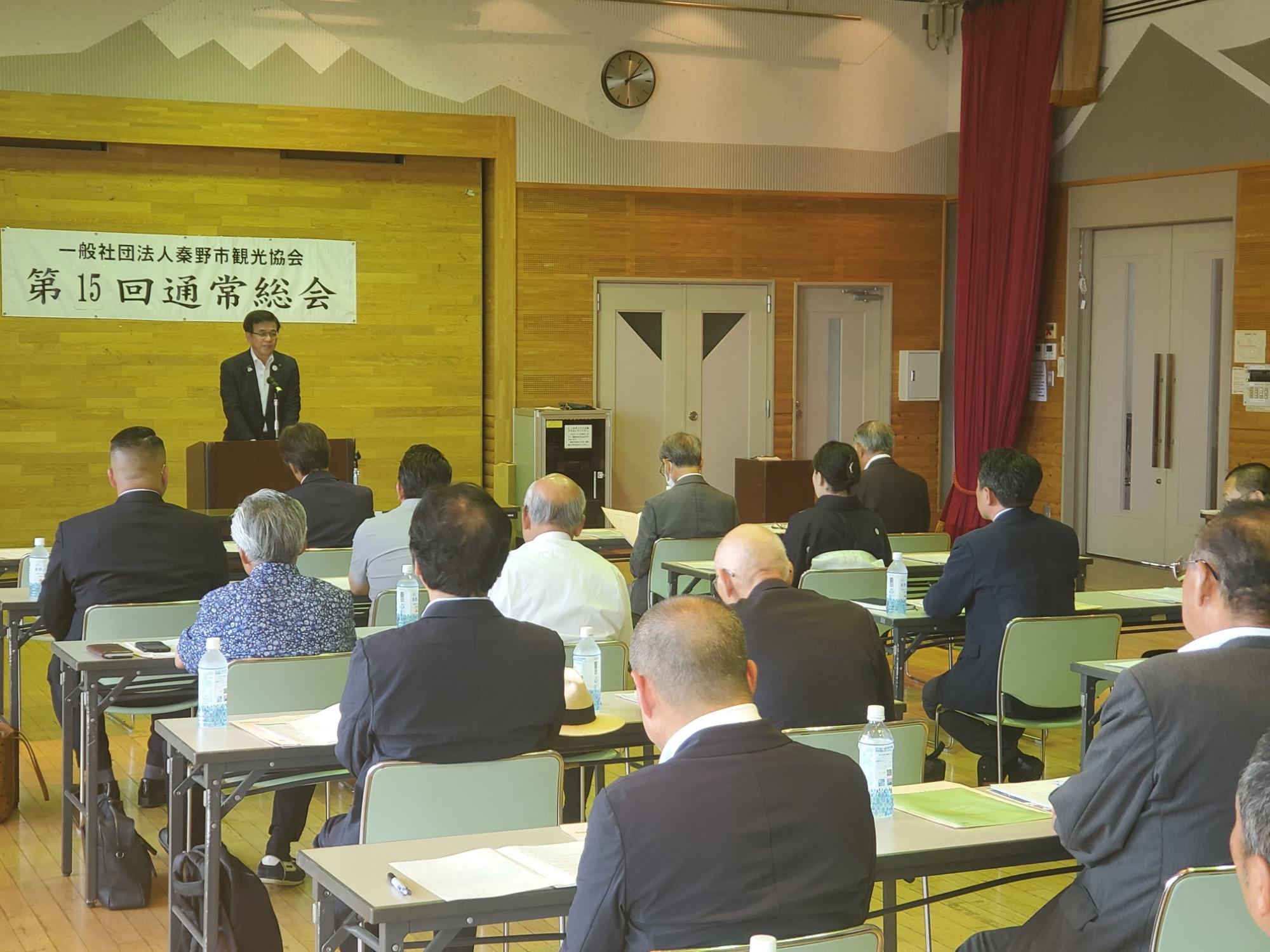 ホールのような会場で、「一般社団法人秦野市観光協会 第15回通常総会」と書かれた横断幕の下、演台に立つ市長が多くの出席者に向かって話しており、聴衆は机と椅子に座って市長を見ている様子の写真