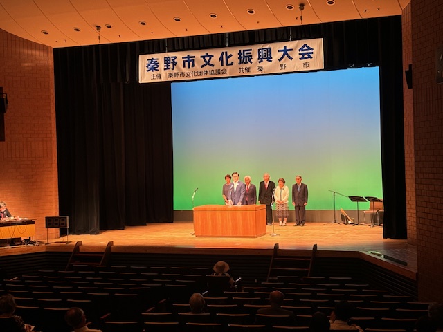 濃い色のカーテンが背景のステージ上で、「秦野市文化振興大会」の横断幕の下に、演台を囲む5人の来賓が立っている写真