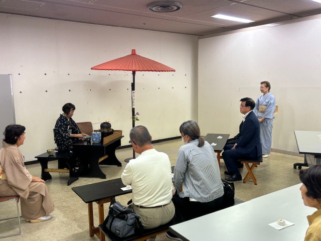 室内に赤い野点傘が立てられ、和服の女性が亭主となり、複数の参加者が茶道体験をしている写真