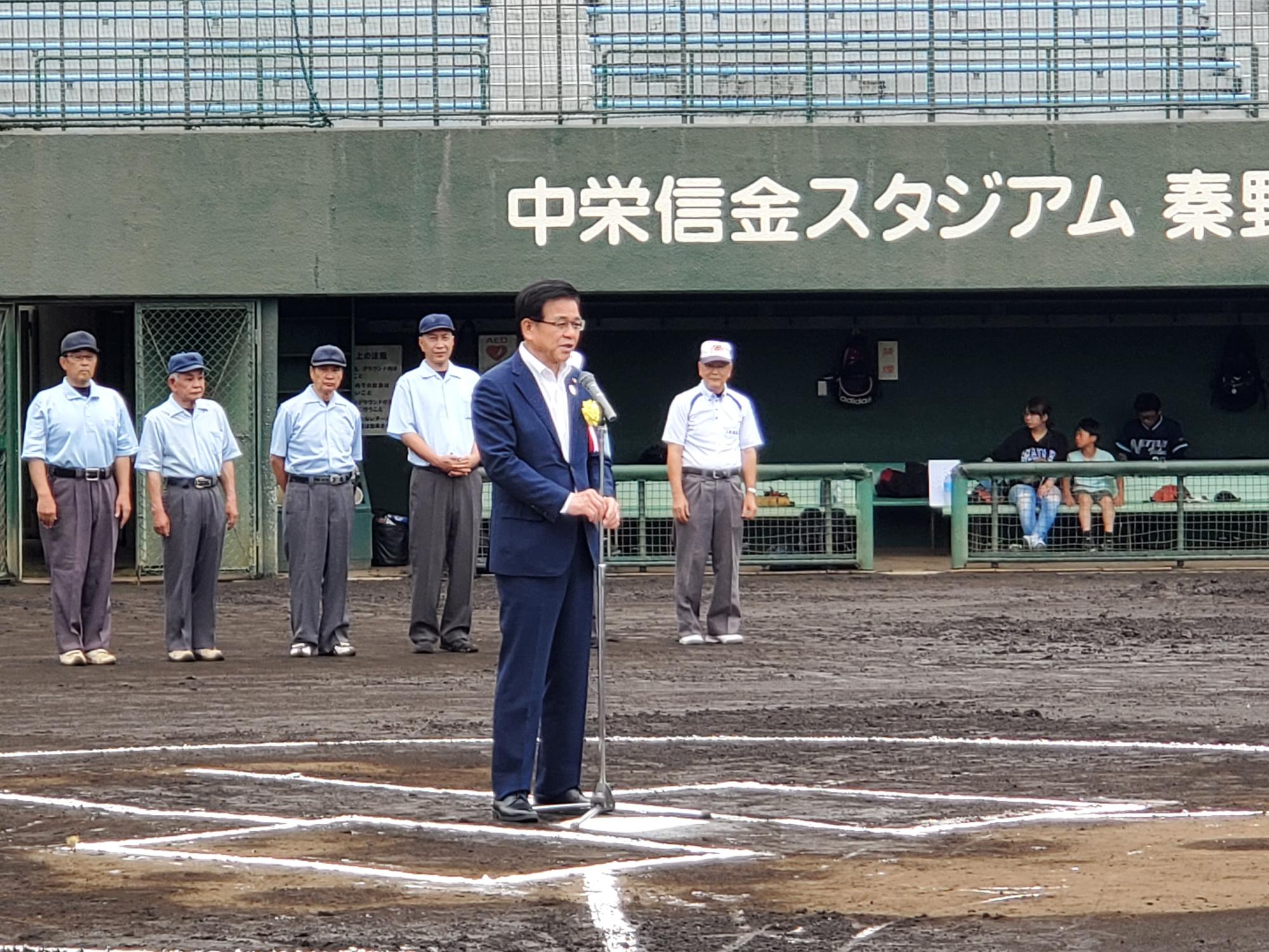 中栄信金スタジアム秦野の土のグラウンドで、スーツ姿の市長がマイクを使って挨拶をしており、その後ろに審判のユニフォームを着た審判たちが整列している写真